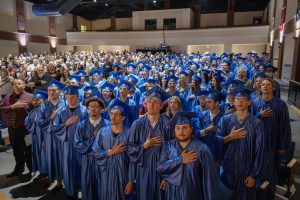 graduates wearing blue caps and gowns stand for the pledge of allegiance