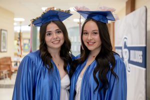 two girls wearing blue caps and gowns pose together in the hallway