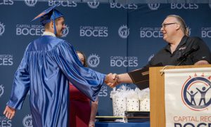 a young man in a blue cap and gown shakes the hand of an adult man at a ceremony