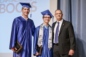 Two young men wearing blue caps and gowns pose next to a man wearing a suit and tie, up on a stage