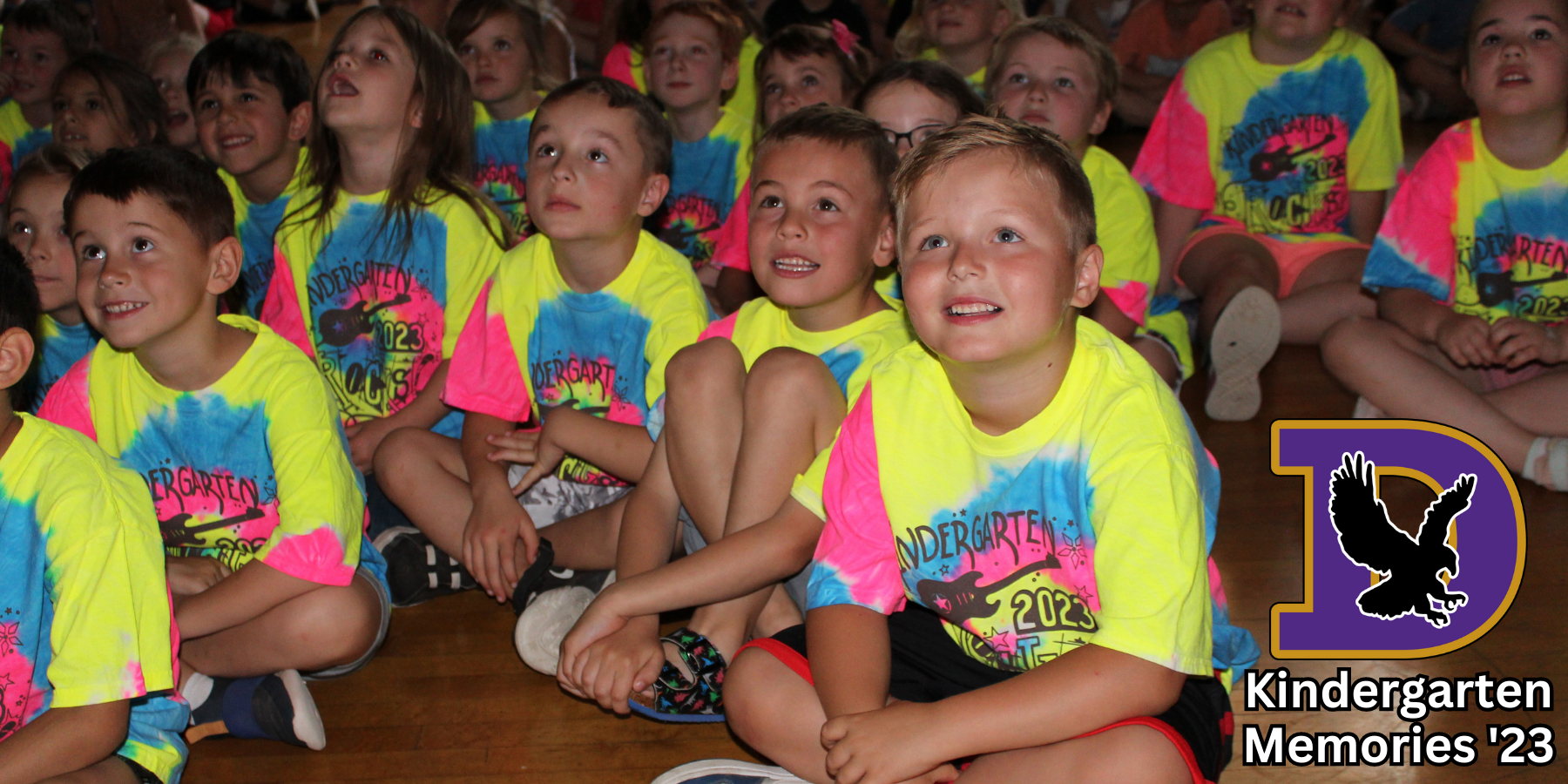 young students wearing tie dye shirts sit on a gym floor watching a movie