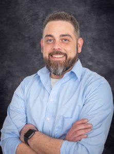 a man with facial hair wears a blue button down shirt and poses with his arms crossed