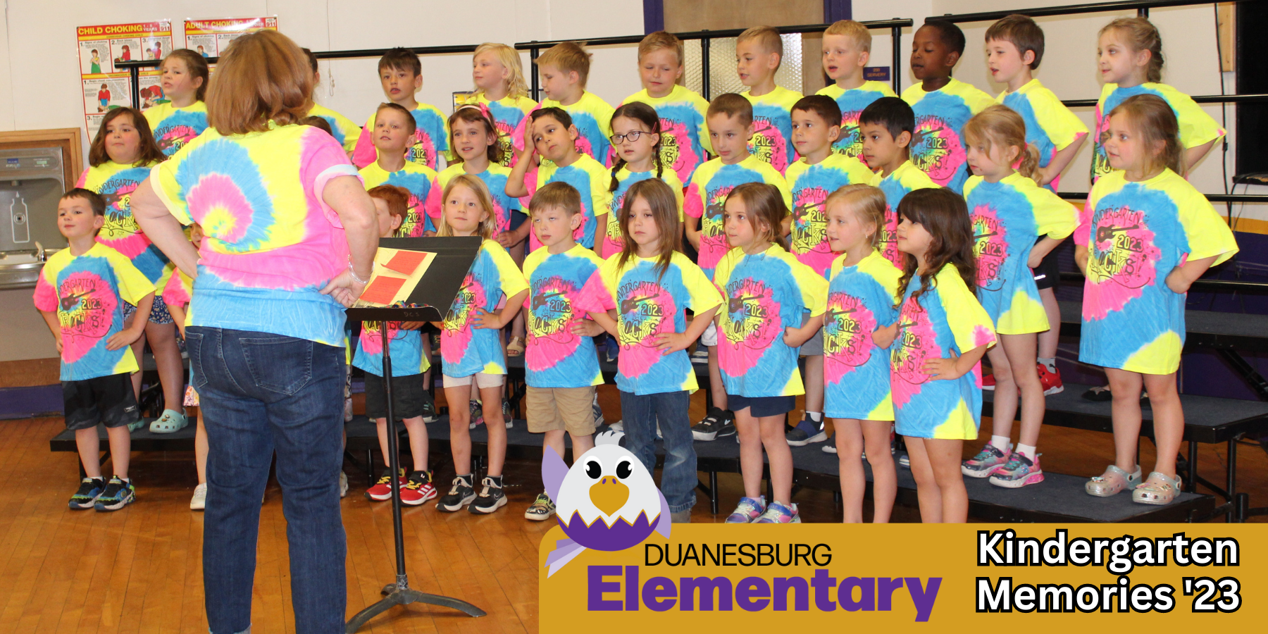 a group of young students wearing tie dye shirts standing on risers, with a logo of an eagle and the words Duanesburg Elementary