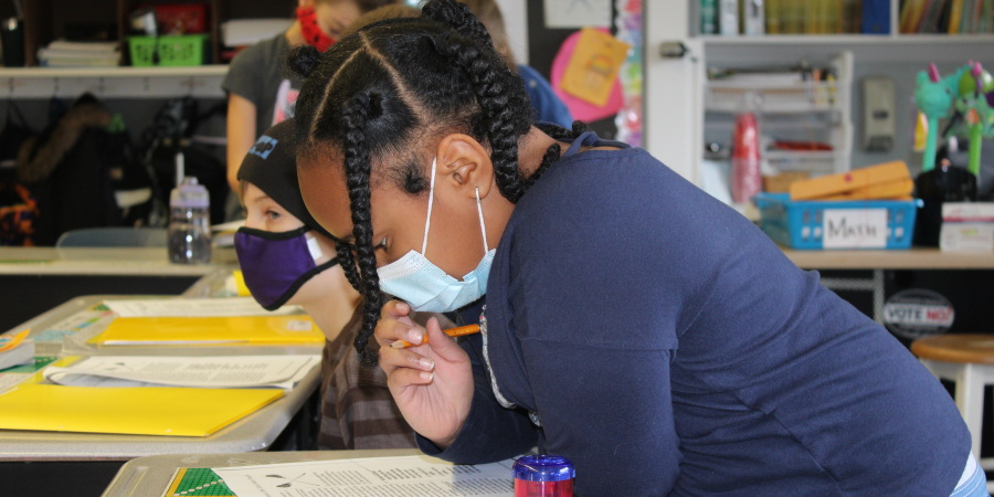 a young girl with a face mask on leans on her desk to complete a homework assignment
