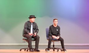 two male actors sit in chairs on a theatrical stage