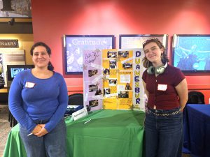 two female students pose next to a table and poster board