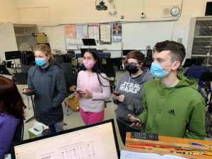 four students wearing face masks are shown looking at a screen that shows a trivia question