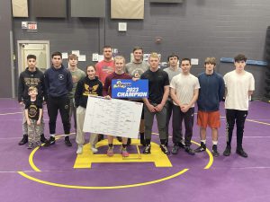 a female wrestler holds up a sign indicating she is a 2023 champion, as other wrestlers stand around her on a purple wrestling mat