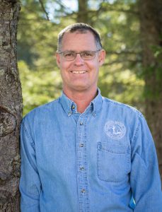 a man wearing glasses and a blue button down shirt poses outside next to a tree