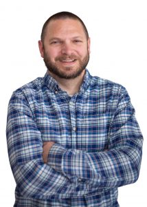 a man wearing a blue plaid shirt and facial hair poses with his arms crossed