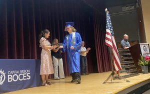 a young man wearing a blue cap and gown receives a diploma on stage