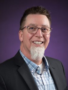 a man wearing glasses, with a white beard, smiles