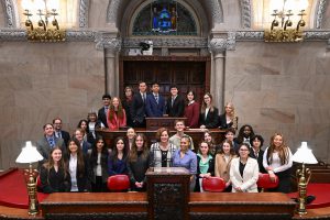 a politician stands in the front and center of a group of students as they pose in the state capitol