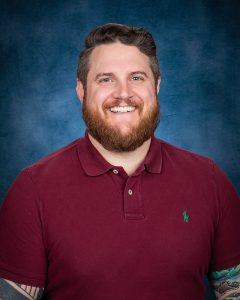 a man with facial hair poses in a short sleeved maroon button down shirt