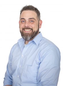 a man wearing a blue button down shirt and facial hair poses in front of a white backdrop