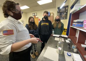 a woman wearing a white shirt and face mask shows three students how to use equipment while on a field trip
