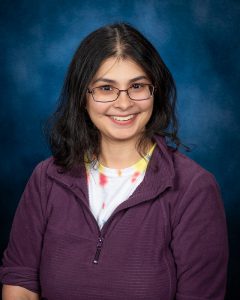 a woman with dark hair and glasses poses in a tie dyed shirt and a maroon fleece jacket
