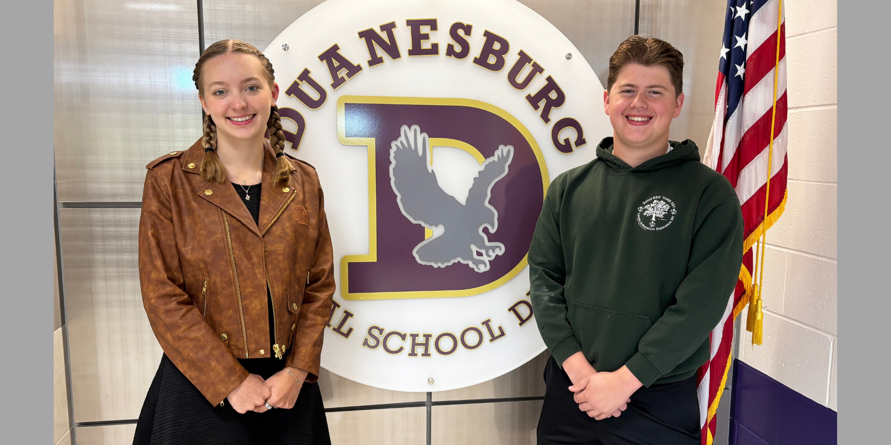two students stand in front of their school logo