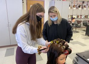 a young girl wearing a face mask puts hair rollers on a model head, as an instructor looks on 