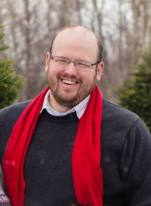 a bald man wearing glasses poses outside in a grey sweater with a red scarf