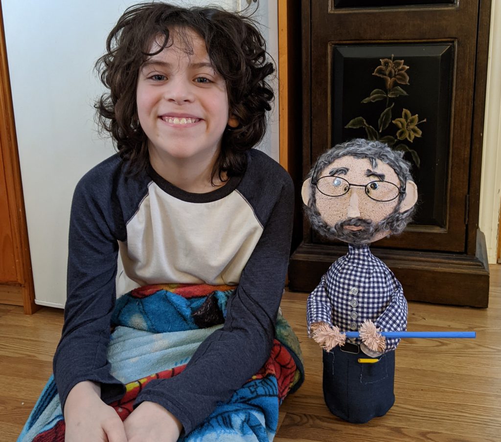 student with curly hair seated next to a 3-d model made out of a soda bottle meant to depict a composer