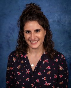 a woman wearing a flowered shirt, with dark hair, poses in front of a blue background