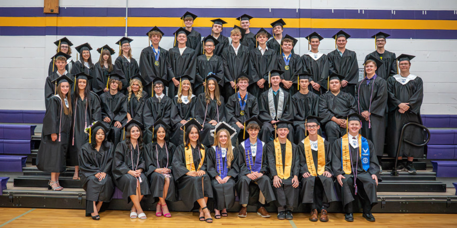 a group of graduates in caps and gowns pose on bleachers