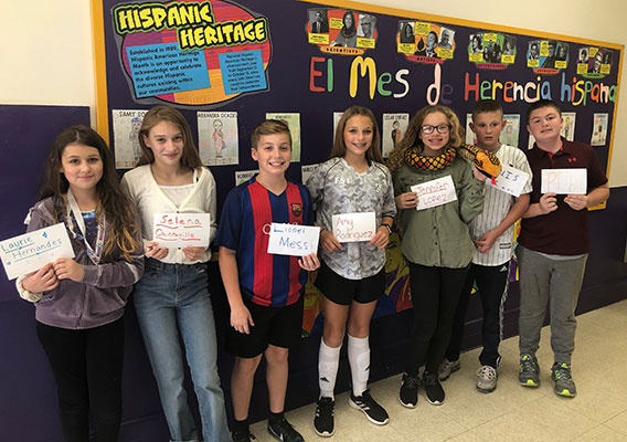 students holding cards with names of Hispanic Americans and standing in front of bulletin board