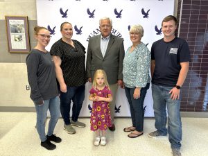 a gentleman with white hair poses in front of a media backdrop with his family and a young girl