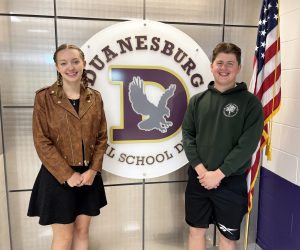 a female and male stand next to each other in front of their school logo and the American flag 