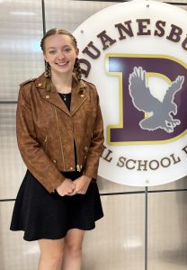 a girl wearing a brown coat and black skirt stands in front of a school logo