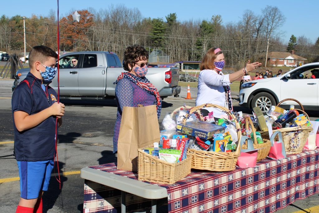 a community member, a student and a teacher stand before a table topped with donated goods in a school parking lot