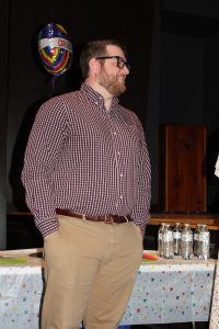 a man with facial hair and glasses poses in an auditorium wearing khaki pants and a checkered shirt 