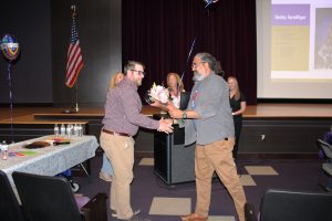 a man wearing khaki pants and a checkered shirt accepts flowers and a balloon from a man with a salt and pepper beard