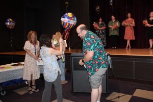 a man wearing a Hawaiin shirt presents flowers and a balloon to a short woman with dark hair