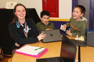 a young adult woman sits at a table with two young elementary students