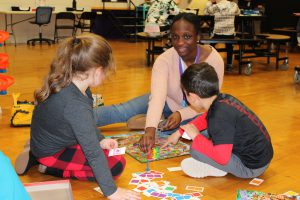 two young students sit on the floor with a teenaged woman and play a board game together