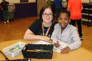 a young woman with glasses sits at a table while a young child colors