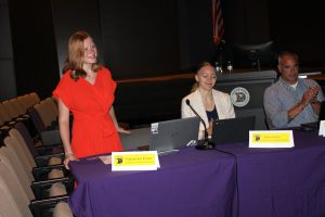 a young girl wearing a red outfit stands behind a purple table, next to another young woman wearing a white blazer sitting down at the table
