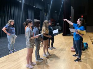 an adult actor stands on a stage with youth actors as they block a scene