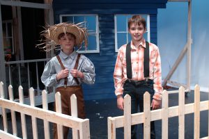 two young boys in plaid shirts and suspenders pose within the set on a theatrical stage