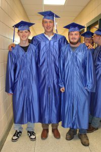 three young men wearing blue caps and gowns pose together in the hallway