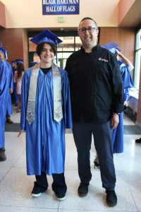 a student wearing a blue cap and gown poses in the hallway next to a chef