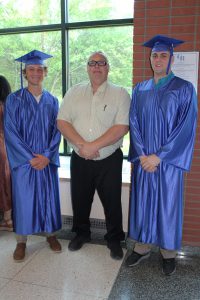 two male students wearing blue caps and gowns pose with their teacher in a hallway