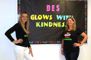 two blonde women wearing black t-shirts stand in front of a bulletin board