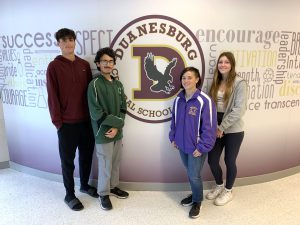 four students - two boys and two girls - pose in a hallway of a school, next to their school's logo