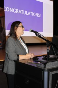 a woman wearing a white shirt and dark jacket with glasses speaks into a microphone