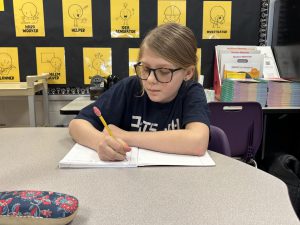 a young girl sits at a table in school and writes a story in a notebook