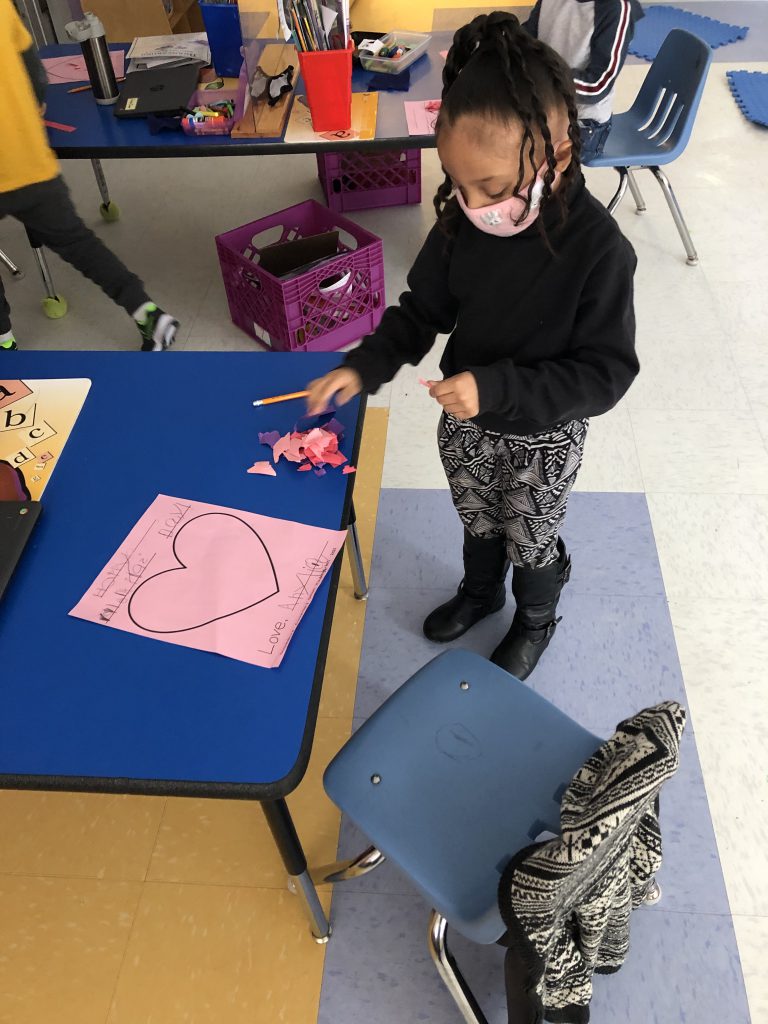 elementary student stands next to a desk while creating a valentine