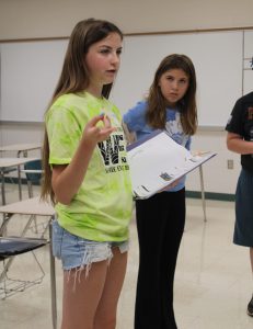 a teenaged girl holds a binder as she addresses a group of students